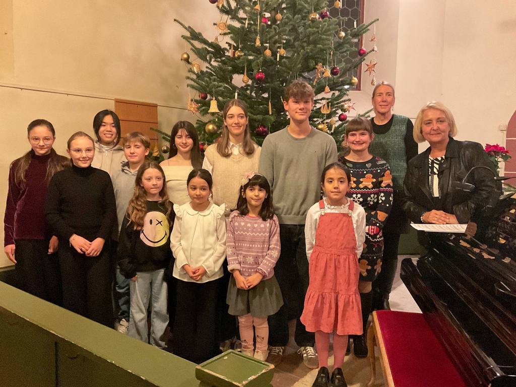 Eine Gruppe von Kindern und Jugendlichen steht in einer Kirche vor einem großen, geschmückten Weihnachtsbaum. Sie tragen festliche Kleidung und lächeln. Rechts im Bild steht eine Frau. Sie hält Notenblätter in der Hand.