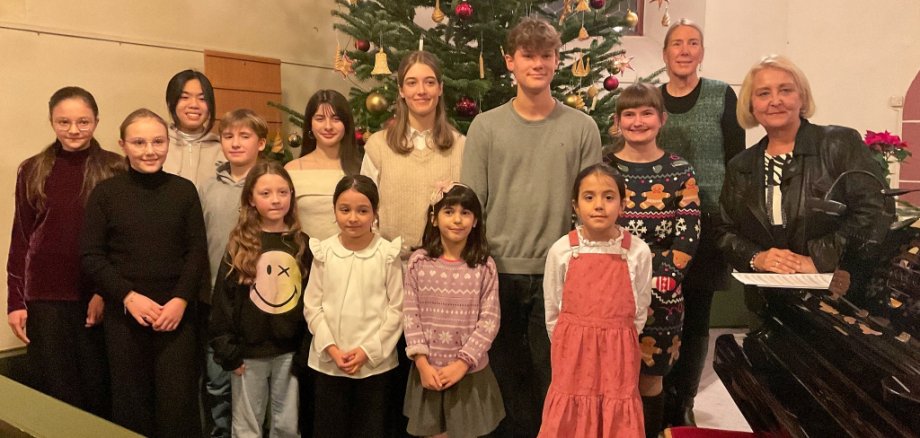 Eine Gruppe von Kindern und Jugendlichen steht in einer Kirche vor einem großen, geschmückten Weihnachtsbaum. Sie tragen festliche Kleidung und lächeln. Rechts im Bild steht eine Frau. Sie hält Notenblätter in der Hand.