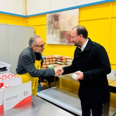 Bürgermeister Dr. Alexander Krey gibt einem älteren Mann mit Brille die Hand. Der ältere Mann trägt eine gelbe Schürze. Im Hintergrund sind Eierkartons und Milchpackungen.
