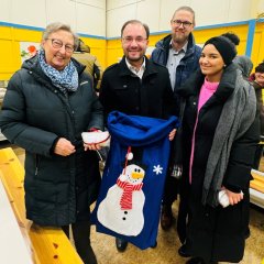 Vier Menschen stehen in einer Halle mit gelben Wänden. Bürgermeister Dr. Alexander Krey hält einen blauen Sack mit einem Schneemann-Motiv. Eine ältere Frau hält einen eingepackten Christstollen. Alle lächeln in die Kamera.