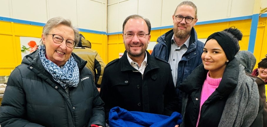 Vier Menschen stehen in einer Halle mit gelben Wänden. Bürgermeister Dr. Alexander Krey hält einen blauen Sack mit einem Schneemann-Motiv. Eine ältere Frau hält einen eingepackten Christstollen. Alle lächeln in die Kamera.