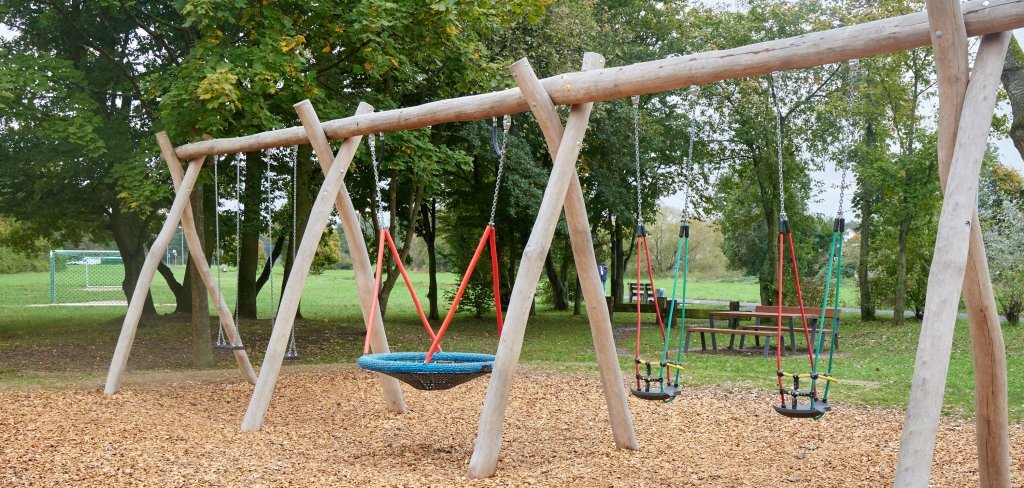 Ein Spielplatz mit mehreren Schaukeln aus Holz. Es gibt eine große Netzschaukel in der Mitte und zwei kleine Schaukeln für Kinder. Der Boden ist mit Holzschnitzel bedeckt. Im Hintergrund stehen Bäume, eine Bank und ein Fußballtor.