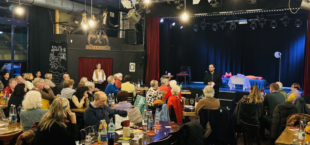 Viele Menschen sitzen an Tischen in einem Saal. Vorne spricht Bürgermeister Dr. Alexander Krey zu den Gästen. Hinter ihm ist eine Bühne mit einem Bett und einer Lampe.