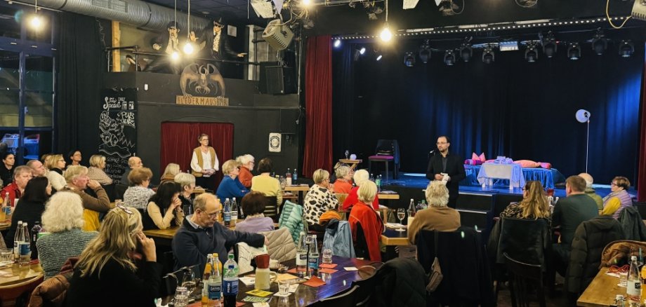 Viele Menschen sitzen an Tischen in einem Saal. Vorne spricht Bürgermeister Dr. Alexander Krey zu den Gästen. Hinter ihm ist eine Bühne mit einem Bett und einer Lampe.