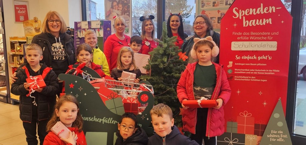 Eine Gruppe von Kindern und Erwachsenen steht in einem Geschäft. Die Kinder halten rote Geschenke in den Händen. Hinter ihnen steht ein kleiner Weihnachtsbaum. Neben dem Bau ist ein großes rotes Schild. Auf dem Schild steht "Spendenbaum". Alle lächeln in die Kamera.