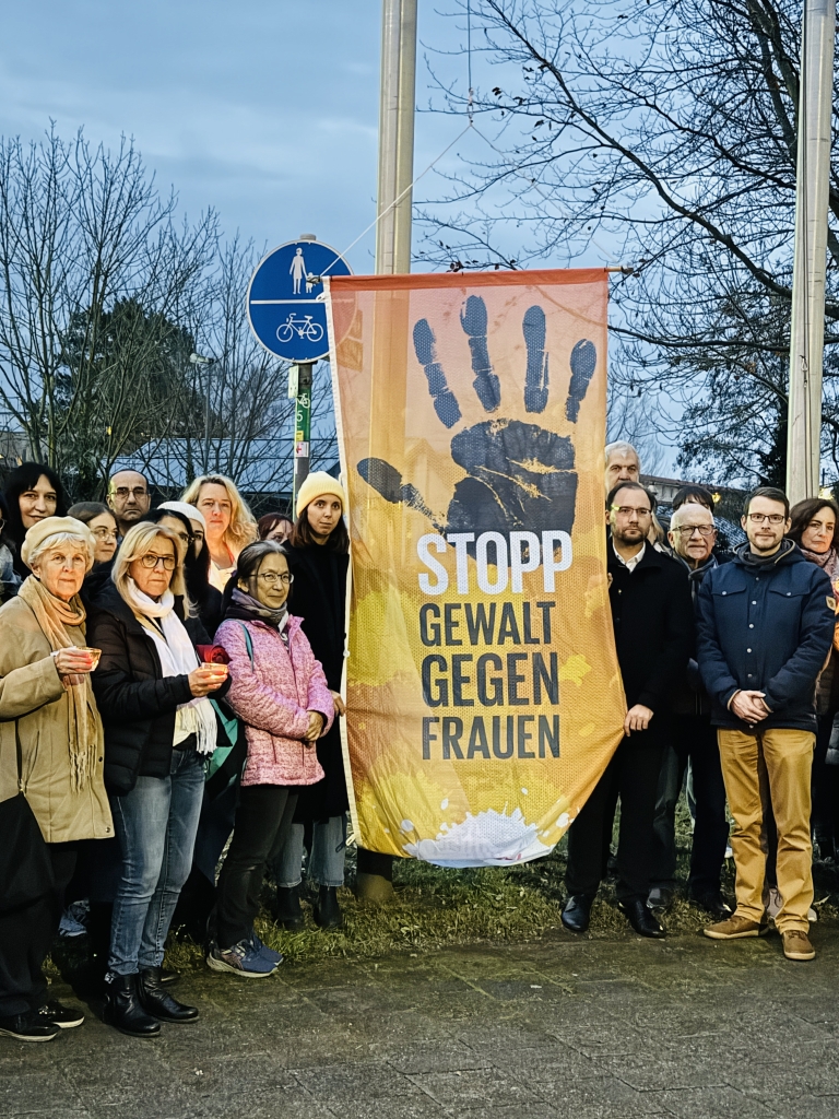 Viele Menschen stehen draußen vor einer Fahne. Auf der Fahne steht: "Stopp Gewalt gegen Frauen". Die Menschen tragen warme Kleidung. Einige halten Kerzen in der Hand.