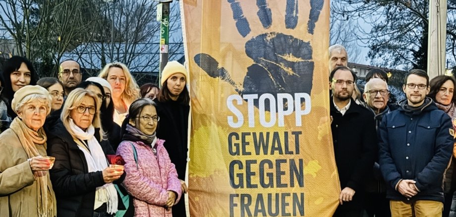 Viele Menschen stehen draußen vor einer Fahne. Auf der Fahne steht: "Stopp Gewalt gegen Frauen". Die Menschen tragen warme Kleidung. Einige halten Kerzen in der Hand.