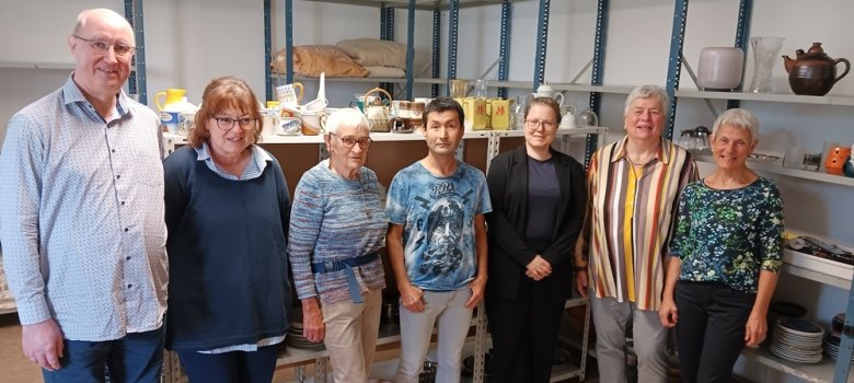 Sieben Menschen stehen vor großen Regalen mit Geschirr und anderen Haushalts-Sachen. Sie helfen mit, dass gespendete Dinge an Menschen weitergegeben werden. Alle schauen freundlich in die Kamera.