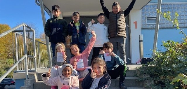 Neun Kinder sitzen und stehen auf einer Treppe vor einem Schwimmbad. Sie lachen und zeigen Daumen nach oben. Einige halten kleine Urkunden in der Hand. Die Sonne scheint. Die Kinder sehen glücklich aus.