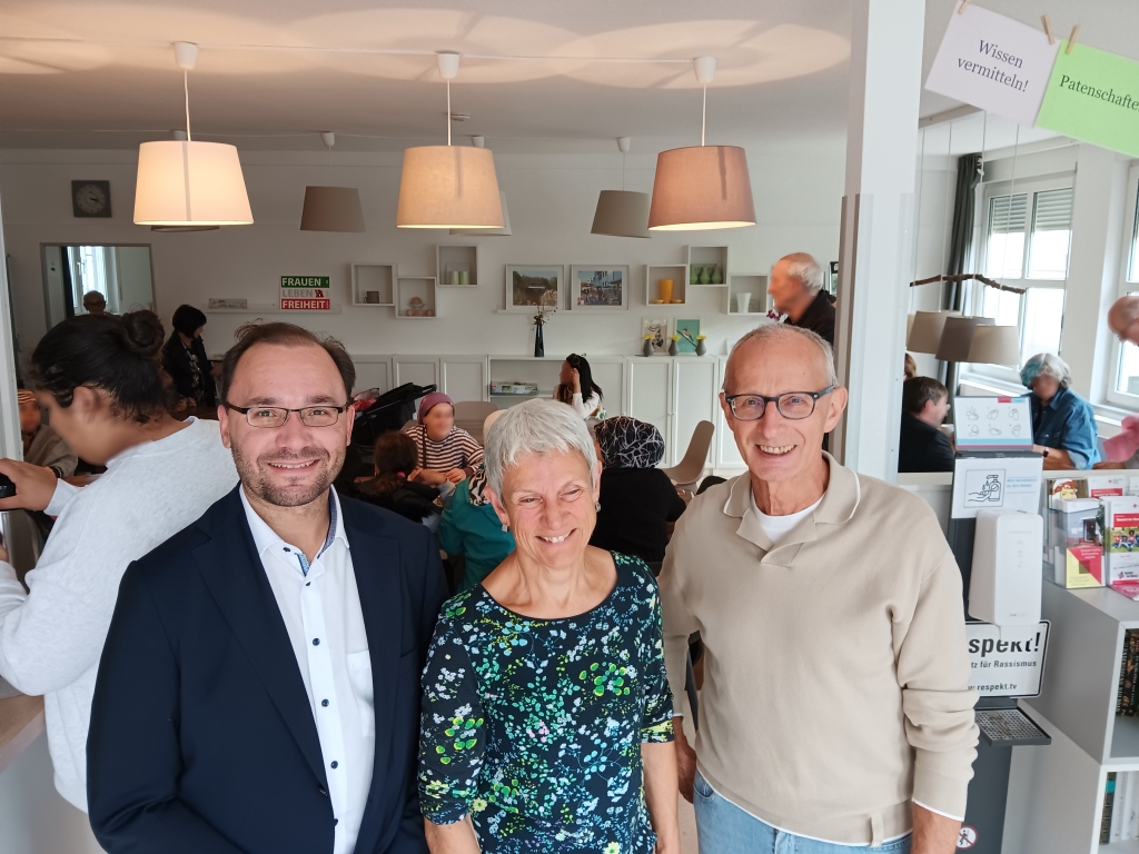 Drei Personen stehen in einem Raum und lächeln. Einer von ihnen ist Bürgermeister Dr. Alexander Krey. Im Hintergrund sitzen Menschen an Tischen und unterhalten sich. Der Raum ist hell und gemütlich.