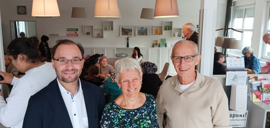 Drei Personen stehen in einem Raum und lächeln. Einer von ihnen ist Bürgermeister Dr. Alexander Krey. Im Hintergrund sitzen Menschen an Tischen und unterhalten sich. Der Raum ist hell und gemütlich.