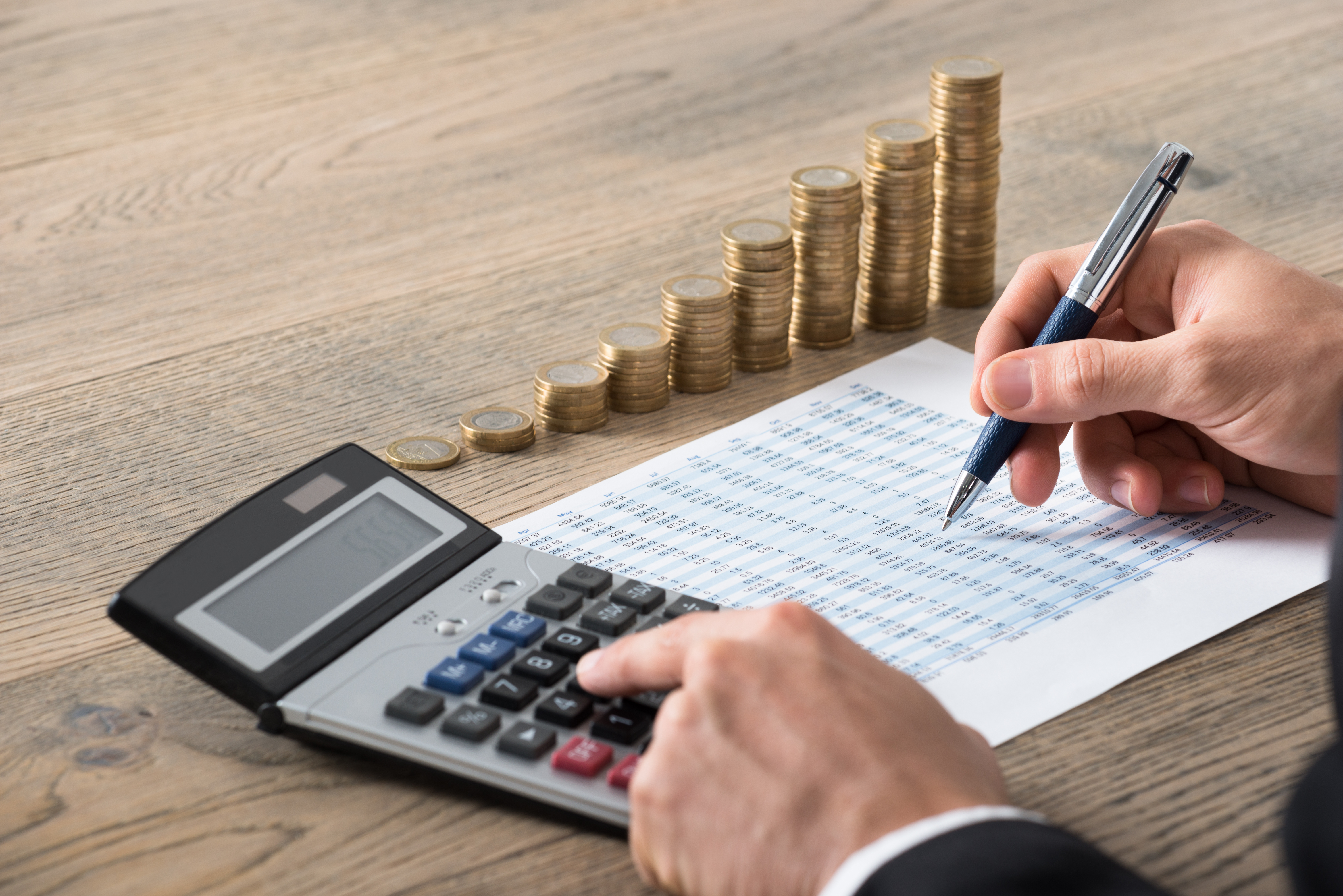Finanzplanung Das Bild zeigt eine Hand mit einem Stift, die auf einem Blatt Papier schreibt. Auf dem Papier sind viele Zahlen zu sehen. Daneben liegt ein Taschenrechner, den die Person gerade benutzt. Im Hintergrund stehen mehrere Stapel Münzen, die aufeinander gestapelt sind und eine ansteigende Linie bilden. Der Tisch hat eine Holzoptik, und die Person trägt einen dunklen Anzug.