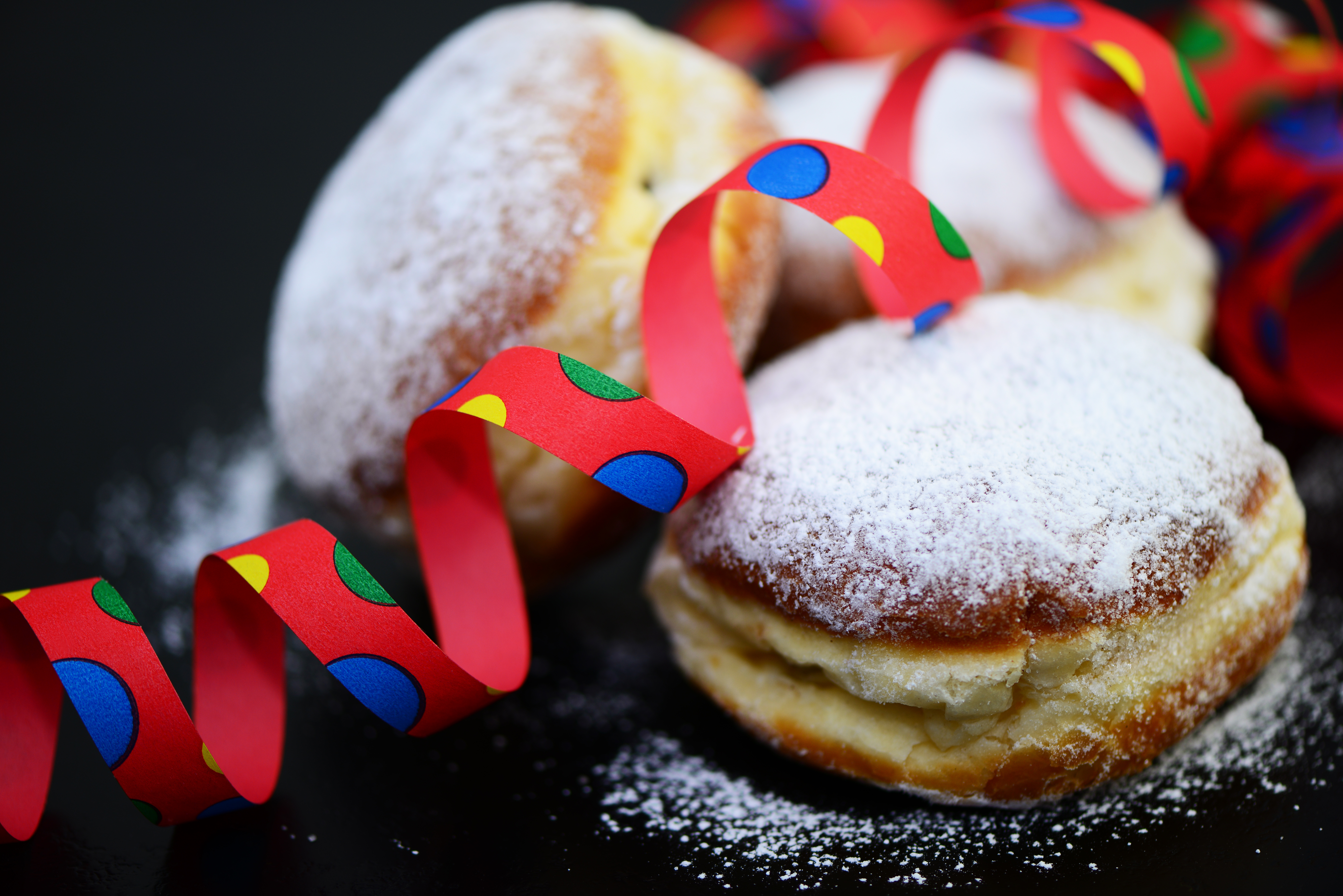 Berliner Das Bild zeigt drei Berliner (Krapfen), die mit Puderzucker bestreut sind. Um die Berliner liegt eine bunte Papierschlange in Rot mit blauen, gelben und grünen Punkten.