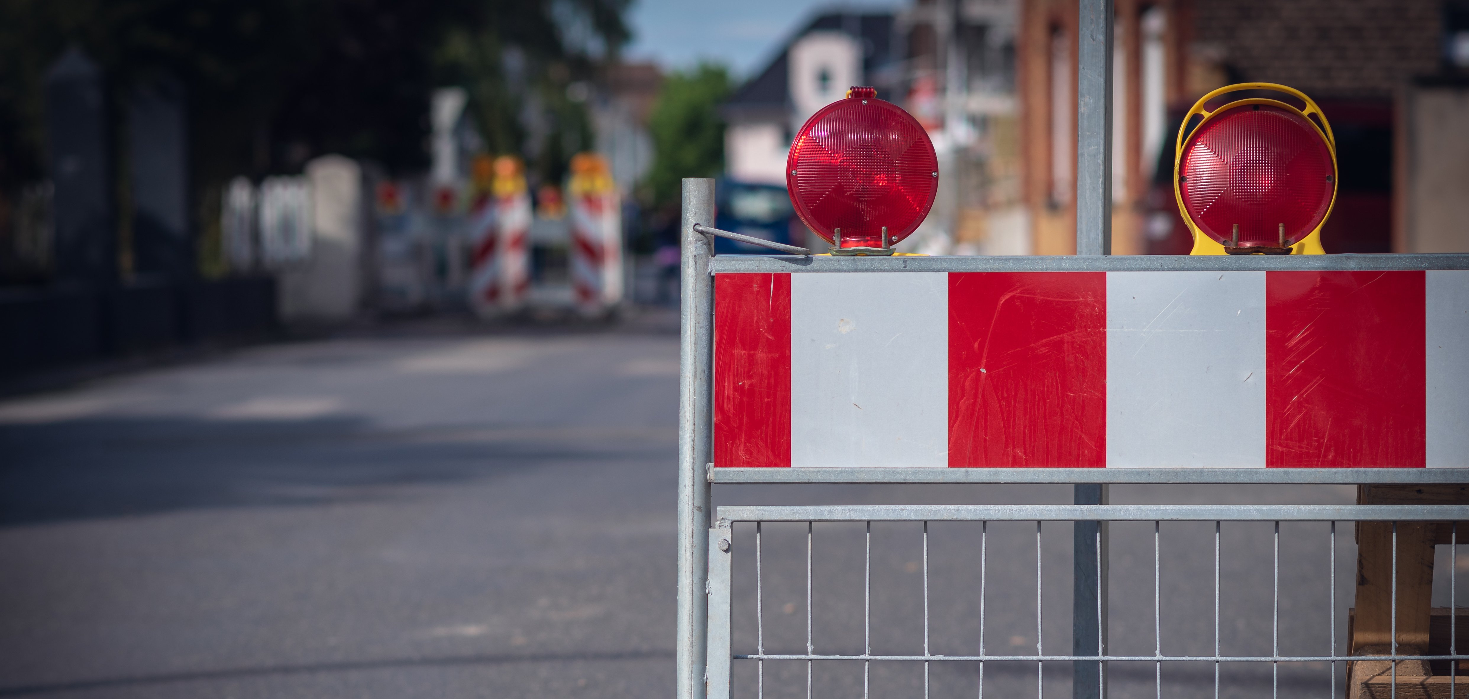 Straßensperrung Auf dem Bild sieht man eine Absperrung auf der Straße. Die Absperrung hat rote und weiße Streifen. Oben sind zwei rote Lampen, die blinken können. Im Hintergrund sieht man weitere Absperrungen und Häuser.