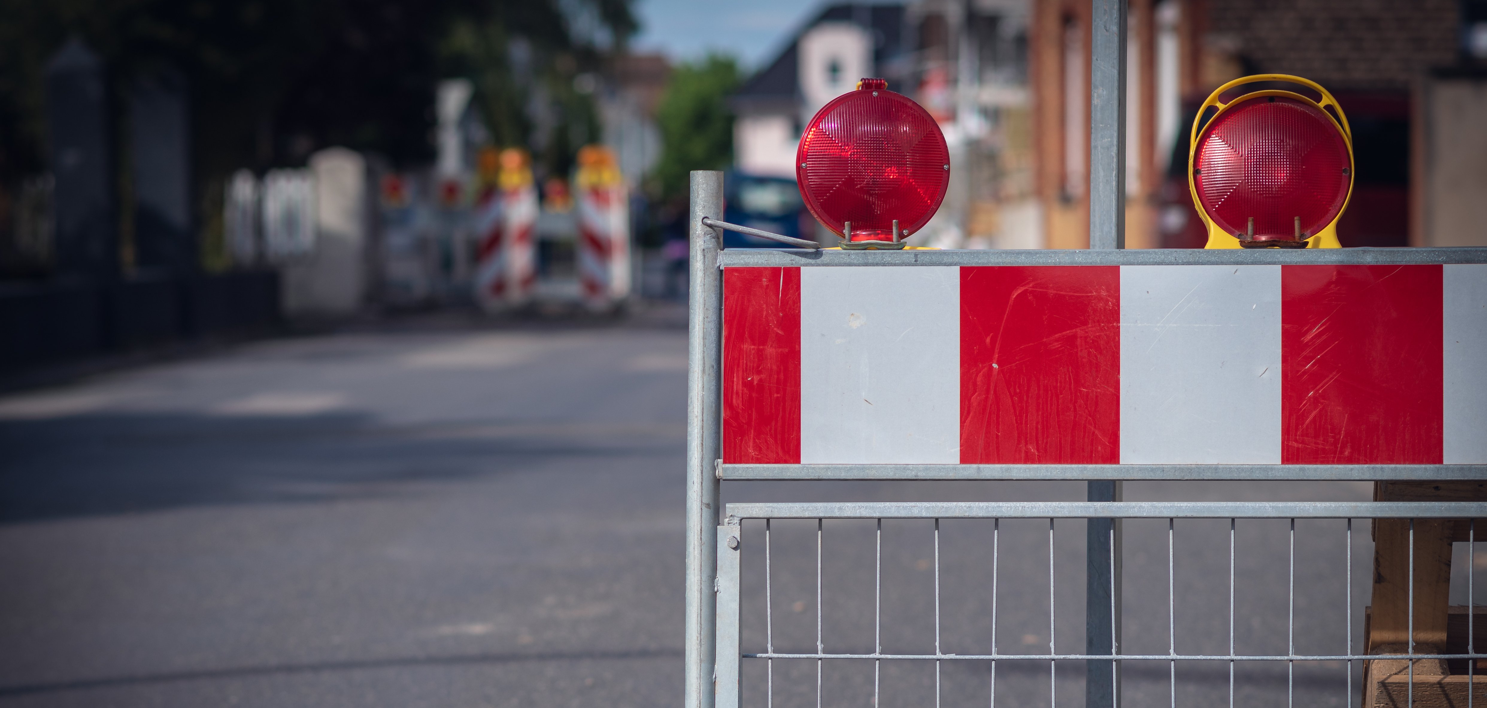 Auf dem Bild sieht man eine Absperrung auf der Straße. Die Absperrung hat rote und weiße Streifen. Oben sind zwei rote Lampen, die blinken können. Im Hintergrund sieht man weitere Absperrungen und Häuser.