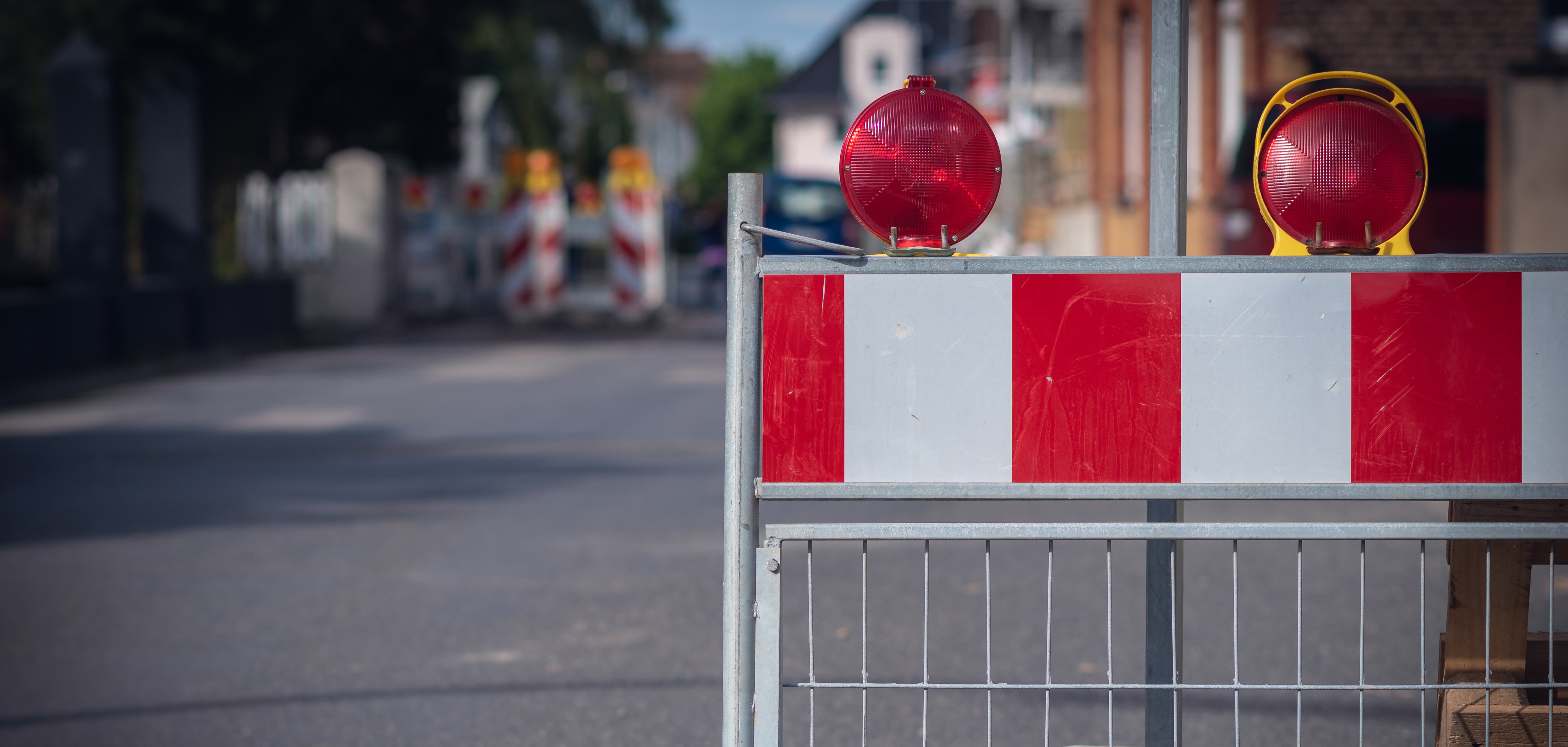 Straßensperrung Auf dem Bild sieht man eine Absperrung auf der Straße. Die Absperrung hat rote und weiße Streifen. Oben sind zwei rote Lampen, die blinken können. Im Hintergrund sieht man weitere Absperrungen und Häuser.