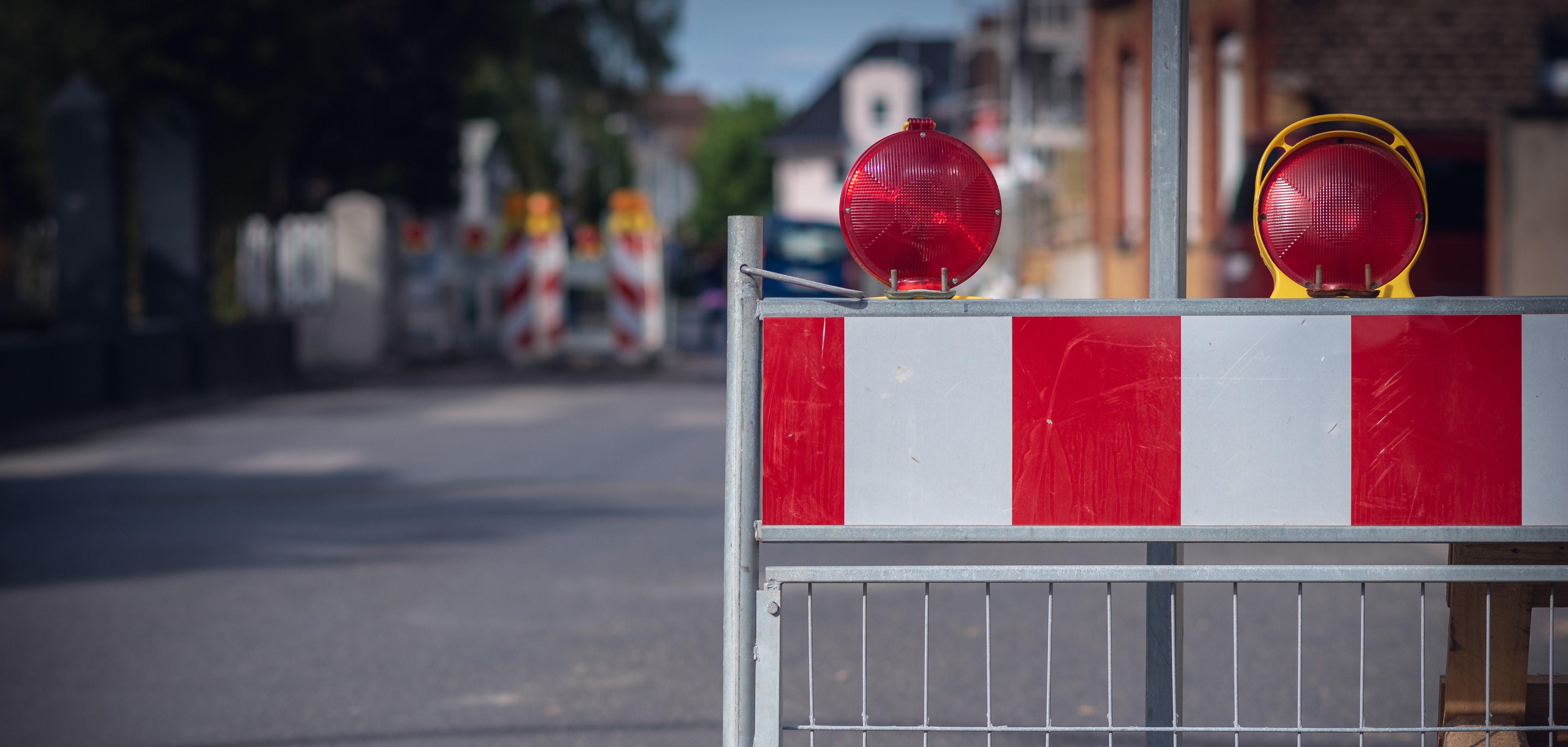 Straßensperrung Auf dem Bild sieht man eine Absperrung auf der Straße. Die Absperrung hat rote und weiße Streifen. Oben sind zwei rote Lampen, die blinken können. Im Hintergrund sieht man weitere Absperrungen und Häuser.