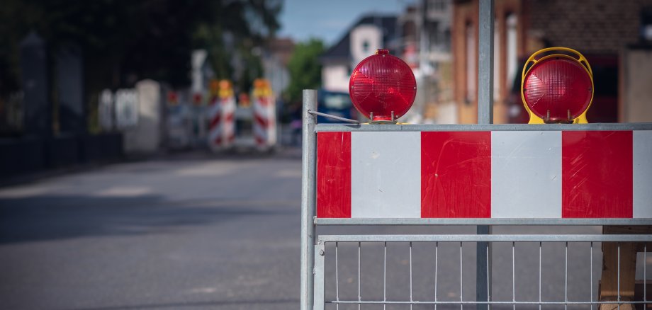 Auf dem Bild sieht man eine Absperrung auf der Straße. Die Absperrung hat rote und weiße Streifen. Oben sind zwei rote Lampen, die blinken können. Im Hintergrund sieht man weitere Absperrungen und Häuser.