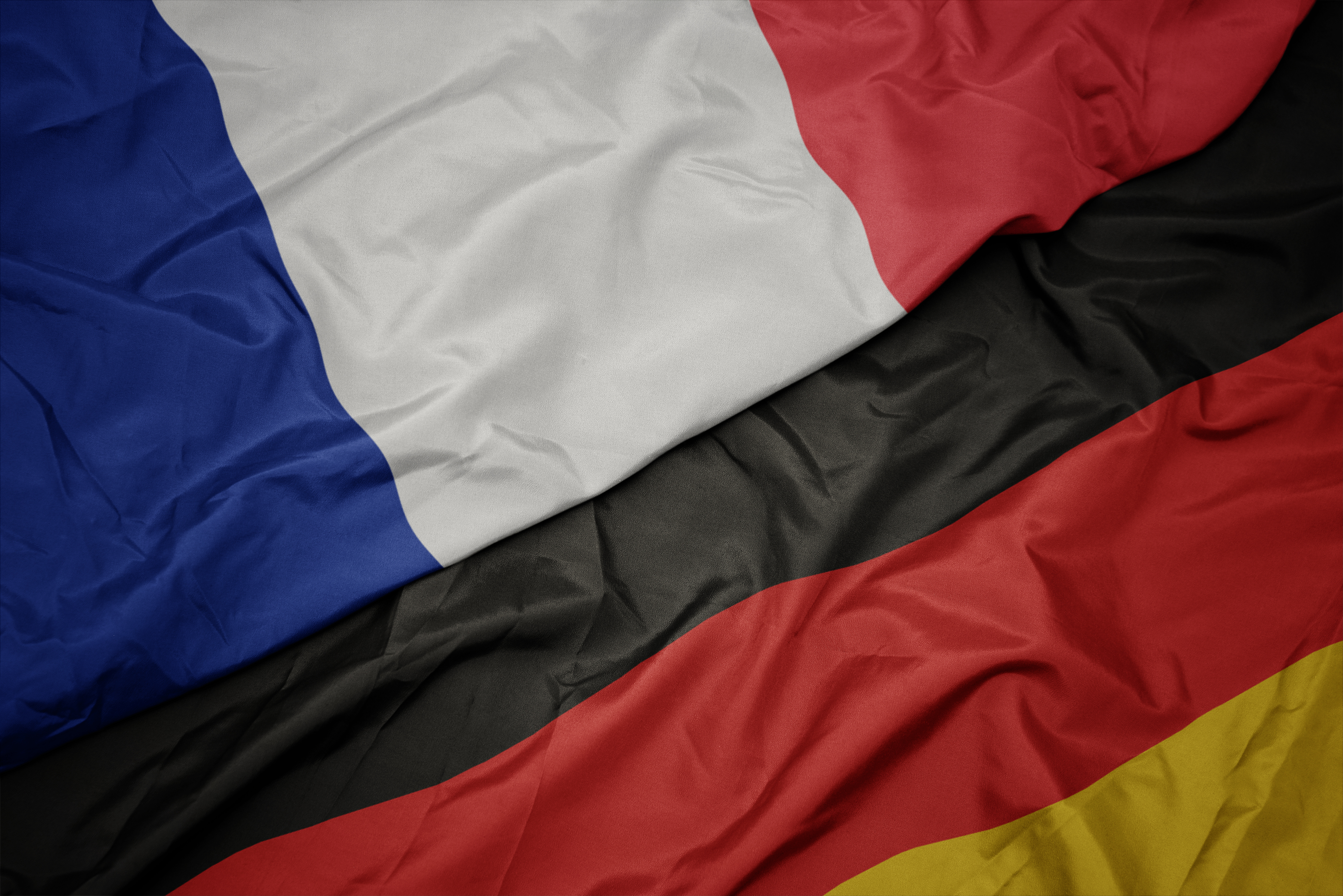 Flaggen von Deutschland und Frankreich Das Bild zeigt zwei Flaggen, die übereinanderliegen. Oben liegt die französische Flagge mit den Farben Blau, Weiß und Rot. Darunter ist die deutsche Flagge in den Farben Schwarz, Rot und Gelb zu sehen. Die Flaggen sind aus Stoff und leicht zerknittert.