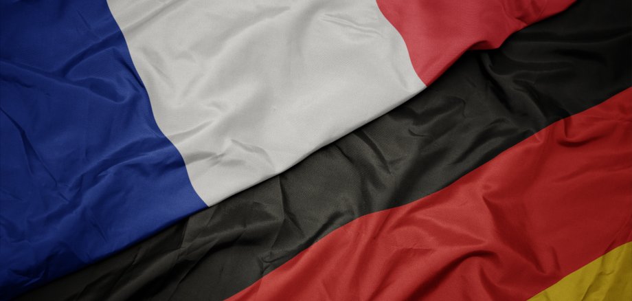Das Bild zeigt zwei Flaggen, die übereinanderliegen. Oben liegt die französische Flagge mit den Farben Blau, Weiß und Rot. Darunter ist die deutsche Flagge in den Farben Schwarz, Rot und Gelb zu sehen. Die Flaggen sind aus Stoff und leicht zerknittert.