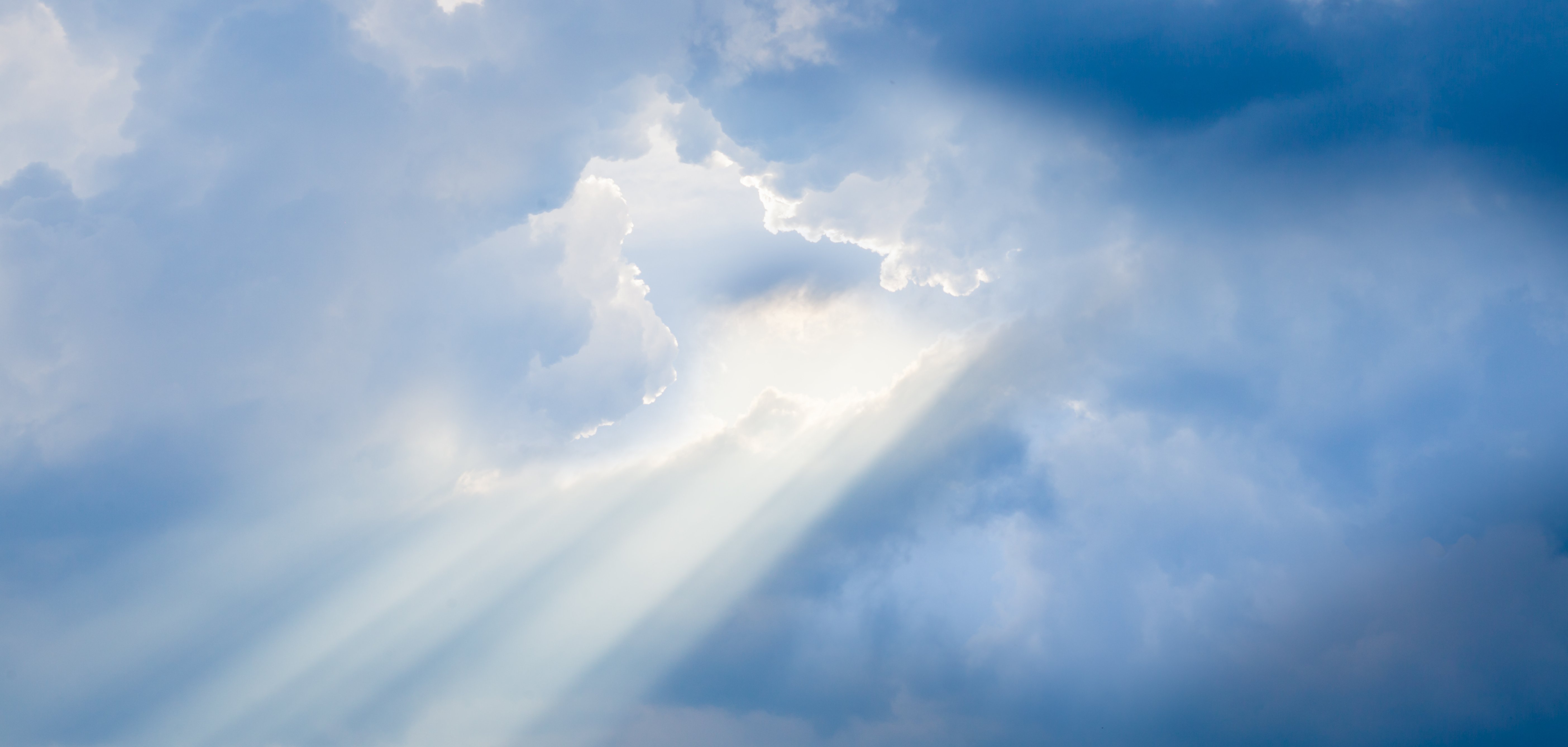 Das Bild zeigt einen Himmel mit vielen weißen und blauen Wolken. Zwischen den Wolken scheint die Sonne hindurch. Sonnenstrahlen brechen durch die Wolkendecke und leuchten hell. Die Lichtstrahlen erzeugen eine friedliche und beruhigende Stimmung.