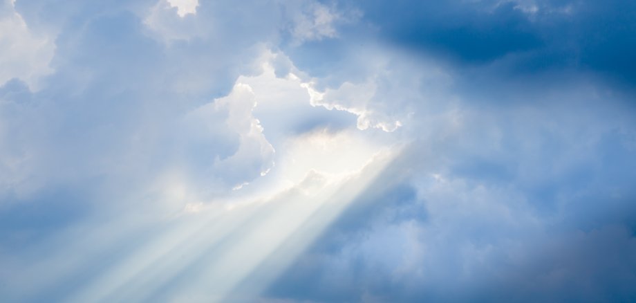 Das Bild zeigt einen Himmel mit vielen weißen und blauen Wolken. Zwischen den Wolken scheint die Sonne hindurch. Sonnenstrahlen brechen durch die Wolkendecke und leuchten hell. Die Lichtstrahlen erzeugen eine friedliche und beruhigende Stimmung.
