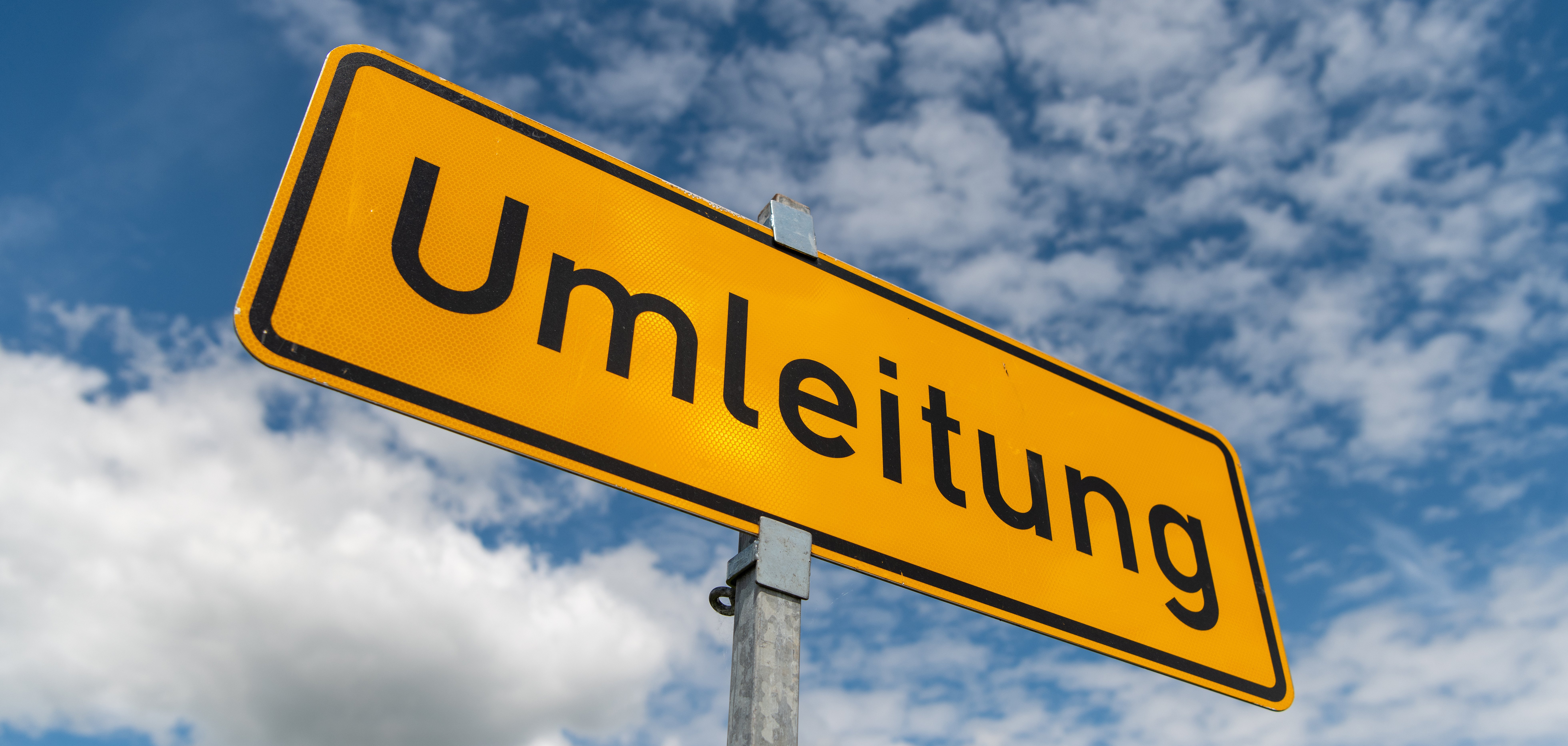 Umleitungsschild Das Bild zeigt ein gelbes Verkehrsschild mit der Aufschrift “Umleitung”. Das Schild hat einen schwarzen Rahmen und schwarze Schrift. Es ist an einem grauen Metallpfosten befestigt. Im Hintergrund ist ein blauer Himmel mit weißen Wolken zu sehen.
