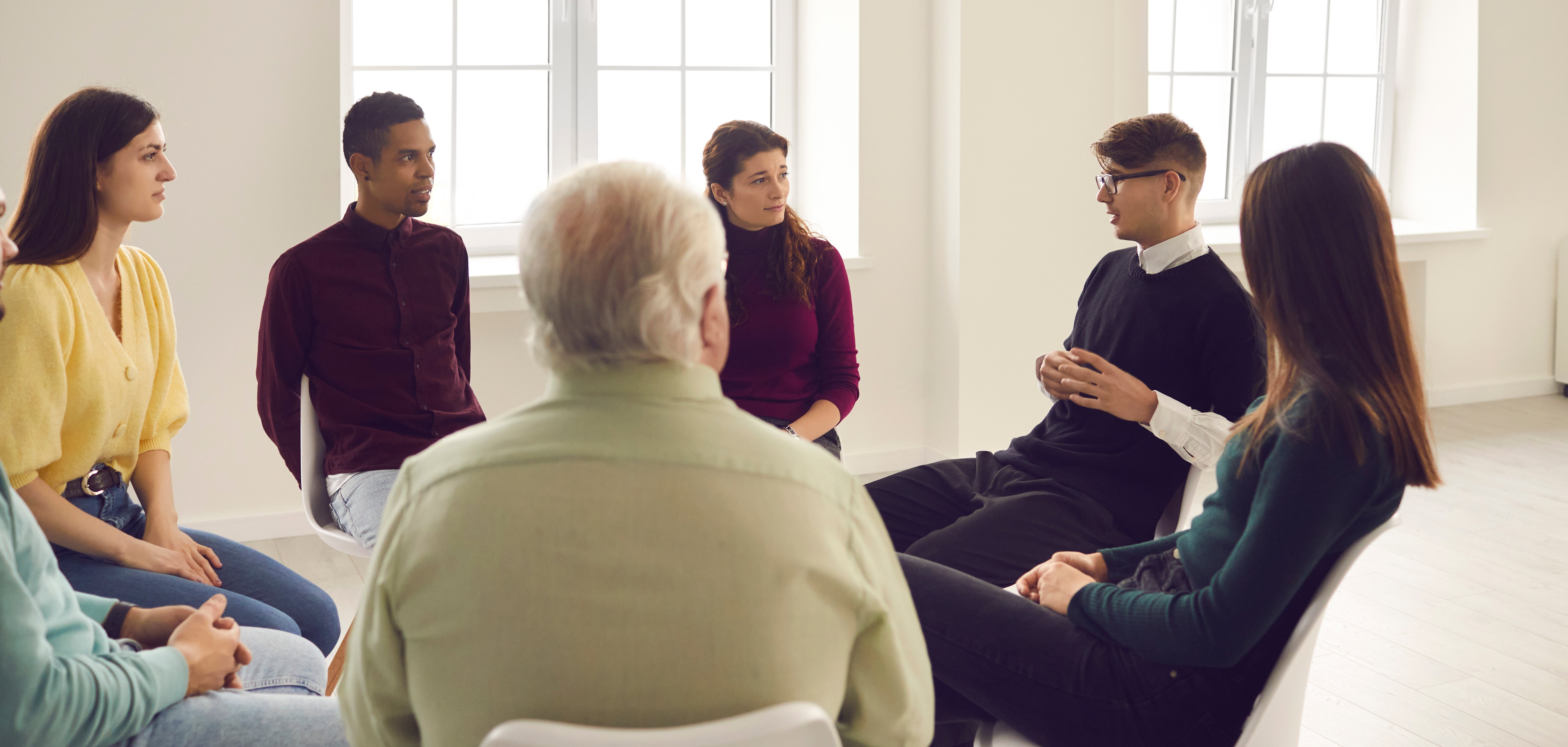 Gruppengespräch Das Bild zeigt eine Gruppe von sieben Personen, die in einem hellen Raum in einem Stuhlkreis sitzen. Die Menschen sind unterschiedlichen Alters und Geschlechts und blicken aufmerksam auf einen jungen Mann in schwarzem Pullover und Brille, der gerade spricht. Der Raum ist schlicht gestaltet, mit großen Fenstern, die viel Tageslicht hereinlassen.