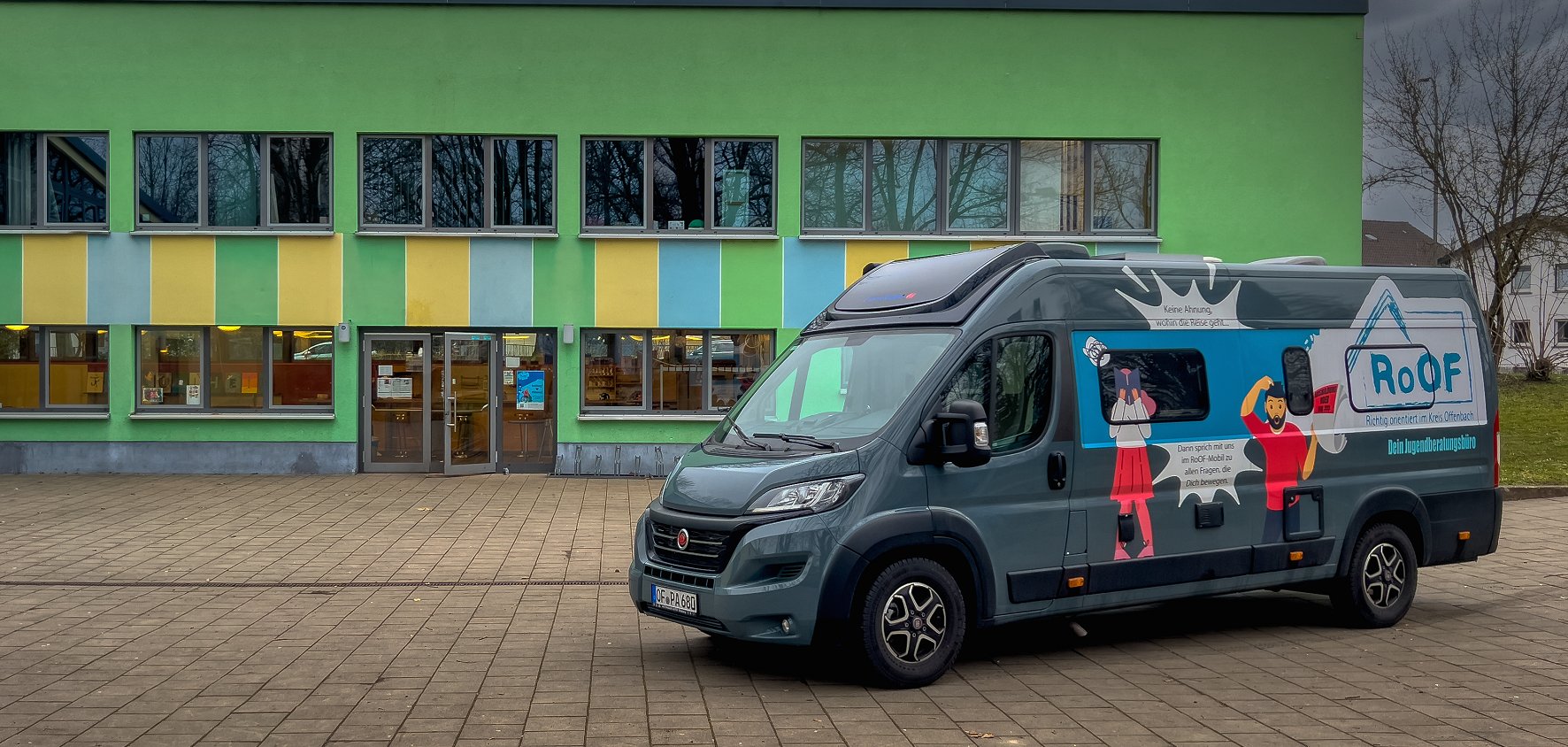 Das RoOF-Mobil vor dem JUZ in Mühlheim Das Bild zeigt ein modernes, grün gestrichenes Gebäude mit großen Fenstern, die teilweise mit gelben und blauen Elementen verziert sind. Vor dem Gebäude steht ein dunkelgrauer Bus mit auffälliger Beklebung. Auf der Seite des Busses sind bunte Illustrationen von Personen sowie das Logo “RoOF” zu sehen. Der Himmel ist bewölkt und grau. Die Bäume im Hintergrund sind kahl. Der Vorplatz ist gepflastert und bietet viel freien Raum vor dem Eingang des Gebäudes.