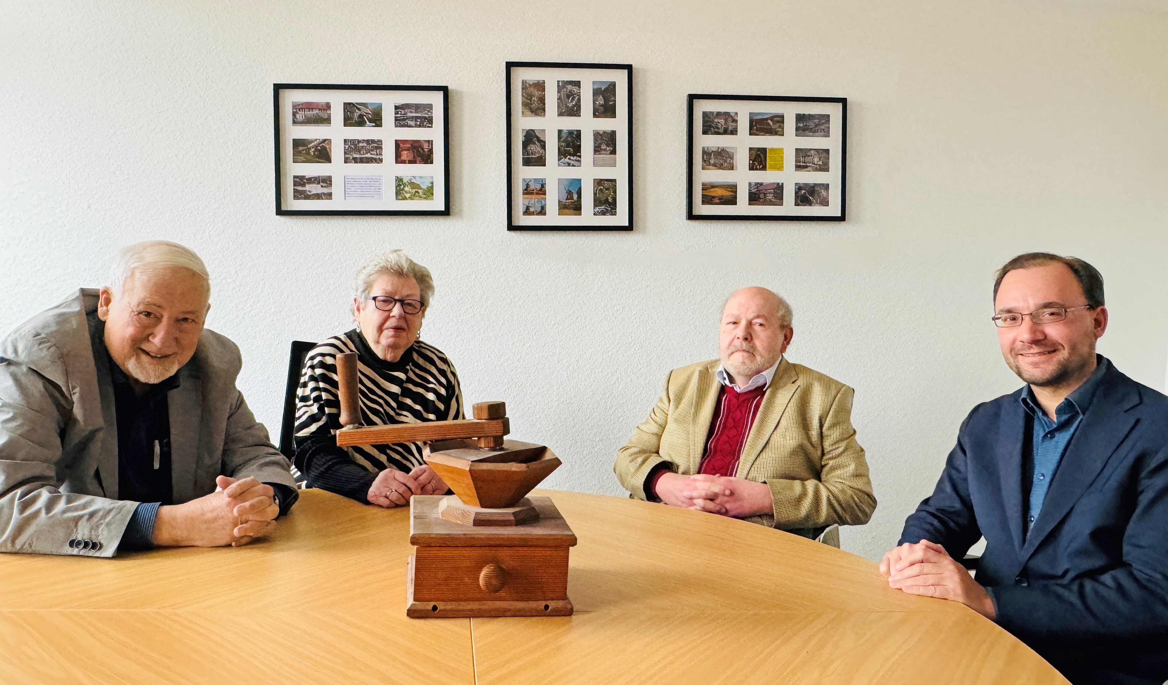 Einweihung Mühlenzimmer Das Bild zeigt vier Menschen an einem runden Holztisch, darunter auch Bürgermeister Dr. Alexander Krey. Auf dem Tisch steht ein hölzernes Objekt, das an eine kleine Mühle erinnert. Im Hintergrund hängen Bilderrahmen mit vielen Fotos. Der Raum erinnert an die Mühlengeschichte der Stadt.