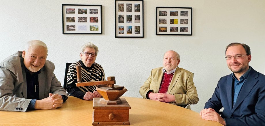 Das Bild zeigt vier Menschen an einem runden Holztisch, darunter auch Bürgermeister Dr. Alexander Krey. Auf dem Tisch steht ein hölzernes Objekt, das an eine kleine Mühle erinnert. Im Hintergrund hängen Bilderrahmen mit vielen Fotos. Der Raum erinnert an die Mühlengeschichte der Stadt.