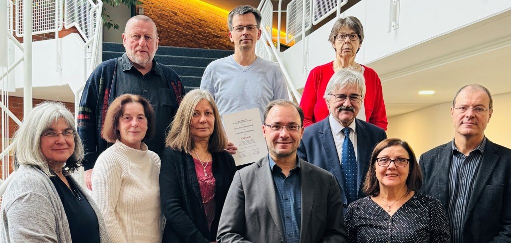 Das Bild zeigt zehn Menschen in einem Gebäude. Sie stehen auf einer Treppe. Einige halten Urkunden in der Hand. Vorne in der Mitte steht der Bürgermeister, Dr. Alexander Krey. Neben ihm steht Rosa Vinci. Sie bekommt eine Urkunde für 25 Jahre Arbeit. Hinter ihnen steht der Erste Stadtrat, Robert Ahrnt. Alle schauen freundlich in die Kamera.