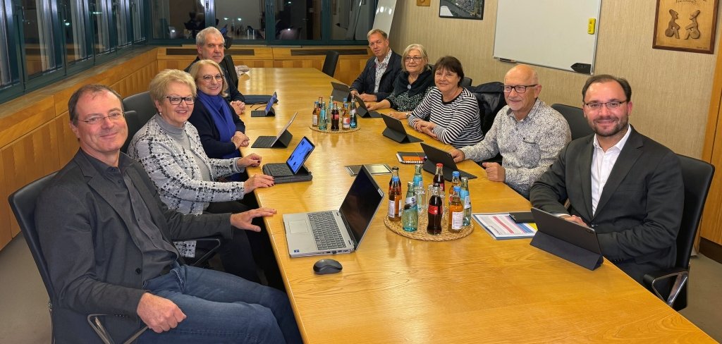 Neun Personen sitzen an einem großen Tisch. Sie haben Laptops und Tablets vor sich. Alle schauen in die Kamera und lächeln. Auf dem Tisch stehen Getränke. Der Raum hat große Fenster und eine weiße Tafel an der Wand.