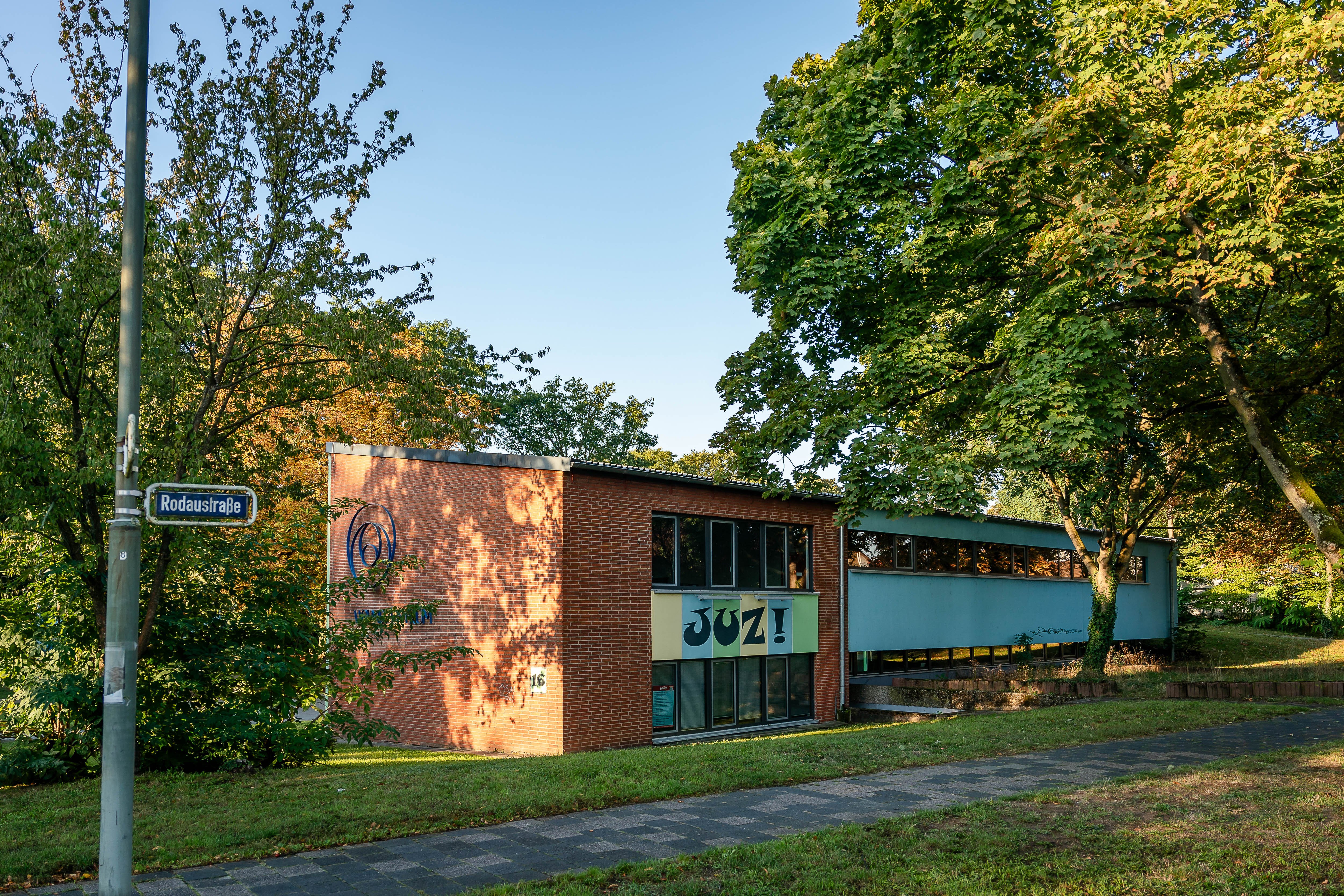 Jugendzentrum Auf dem Bild sieht man ein rotes Backsteingebäude mit großen Fenstern. Auf dem Gebäude steht "JUZ!", das ist die Abkürzung für "Jugendzentrum". Es steht in Mühlheim am Main in der Rodaustraße. Rund um das Gebäude sind viele grüne Bäume. Die Sonne scheint und es wirkt einladend und freundlich.