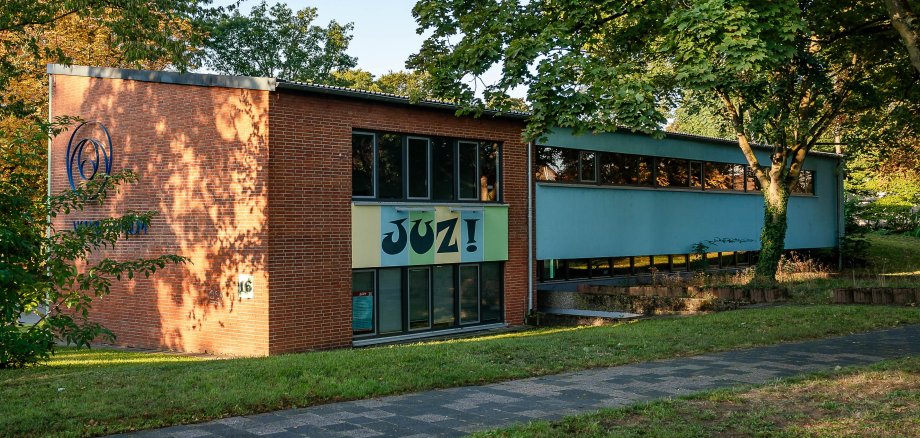 Auf dem Bild sieht man ein rotes Backsteingebäude mit großen Fenstern. Auf dem Gebäude steht "JUZ!", das ist die Abkürzung für "Jugendzentrum". Es steht in Mühlheim am Main in der Rodaustraße. Rund um das Gebäude sind viele grüne Bäume. Die Sonne scheint und es wirkt einladend und freundlich.