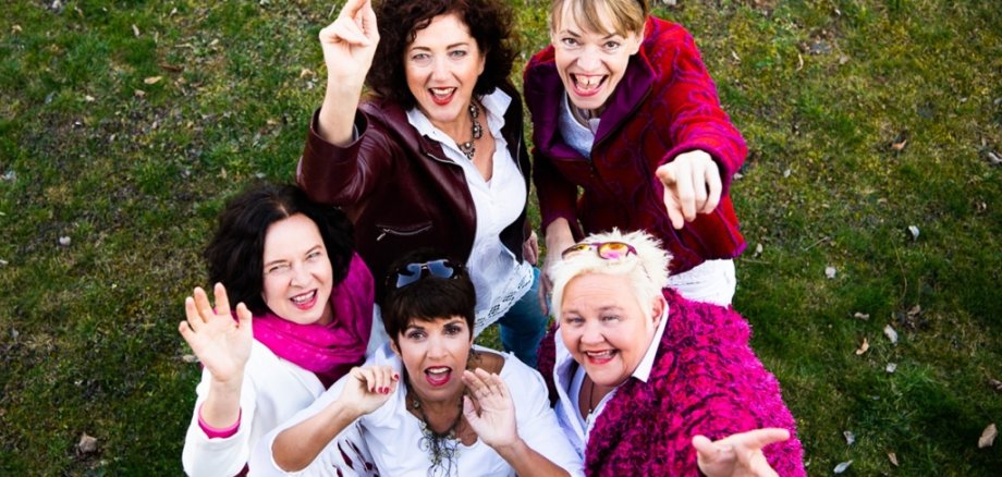 Fünf Frauen stehen auf einer Wiese. Sie schauen lachend nach oben in die Kamera. Sie tragen bunte Kleidung in weiß, pink und rot. Einige zeigen mit den Fingern nach oben oder winken.