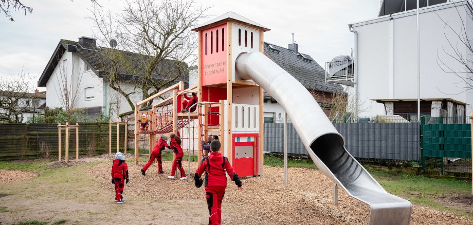 Das Bild zeigt einen Spielplatz mit einem großen Klettergerüst in Form eines roten Feuerwehrturms. Mehrere Kinder in roten Overalls spielen auf dem Gerüst. Einige klettern, andere laufen auf den Spielplatz zu. Das Klettergerüst hat eine lange silberne Rutsche und viele Kletter- und Spielmöglichkeiten. Im Hintergrund sind Wohnhäuser und ein kahler Baum zu sehen. Der Boden ist mit Holzschnitzeln bedeckt.