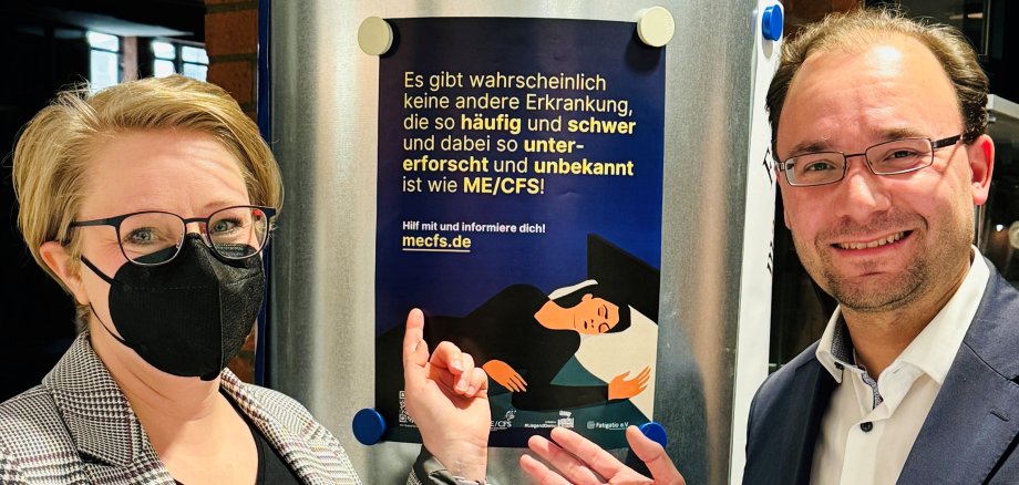 Das Bild zeigt zwei Personen vor einer silbernen Metallsäule. Die Frau links trägt eine schwarze Schutzmaske, eine Brille und ein kariertes Jackett. Der Mann rechts ist Bürgermeister Dr. Alexander Krey. Er trägt ein weißes Hemd und ein dunkles Sakko. Beide lächeln und zeigen mit den Händen auf ein Plakat, das an der Säule befestigt ist. Das Plakat ist dunkelblau mit gelbem Text. Es zeigt eine liegende Person und informiert über die Krankheit ME/CFS. Im Hintergrund sind eine Backsteinwand und ein Fenster mit einfallendem Tageslicht erkennbar.