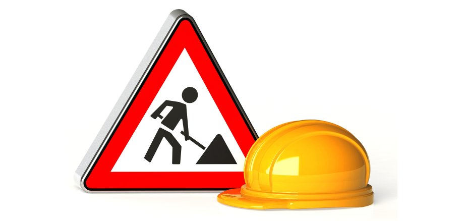 Das Bild zeigt ein dreieckiges Baustellenschild und einen gelben Schutzhelm. Das Schild hat einen roten Rand und ein schwarzes Symbol. Es zeigt eine Person, die mit einer Schaufel arbeitet. Der gelbe Bauhelm liegt schräg vor dem Schild. Der Hintergrund ist weiß und schlicht.