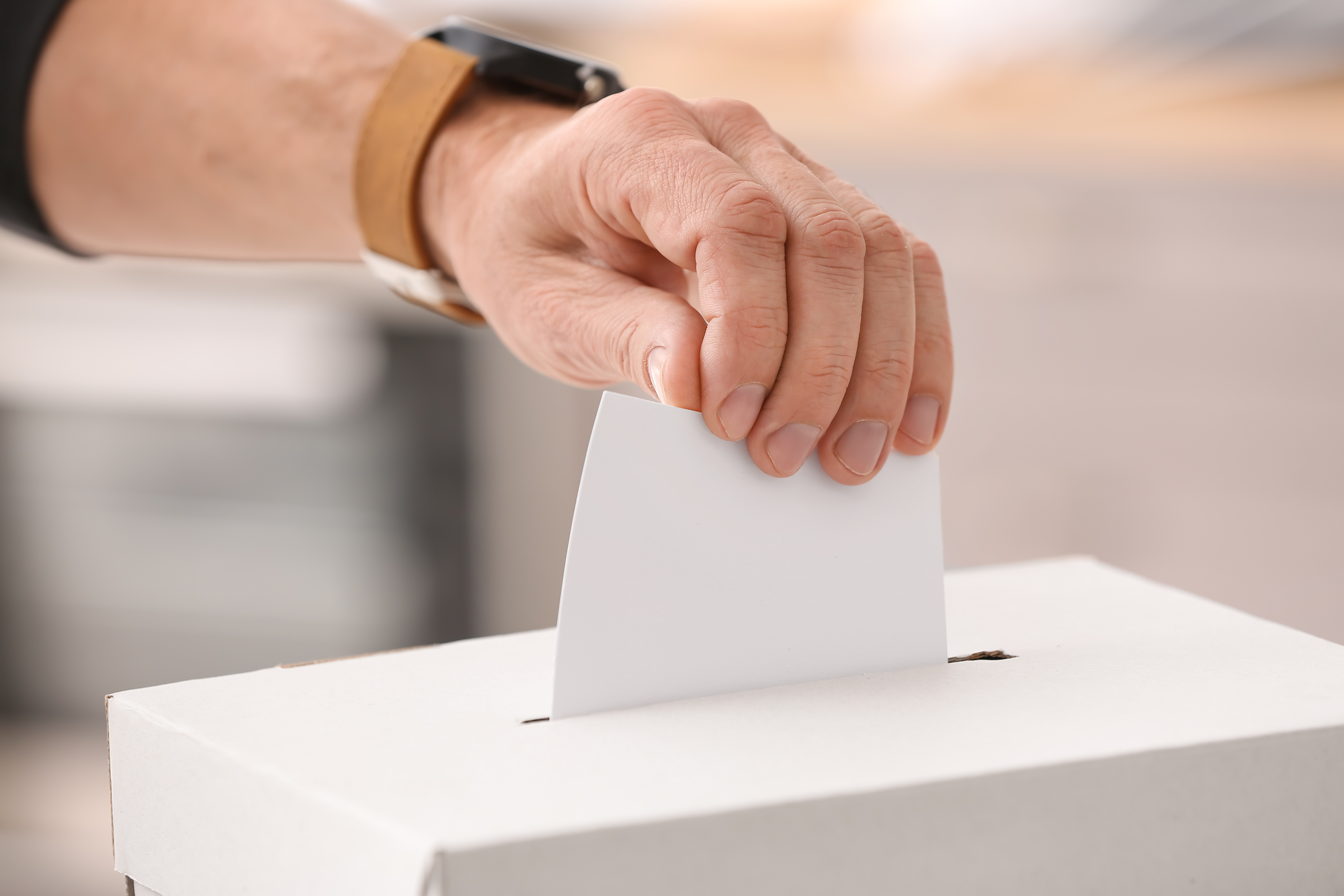 Stimmabgabe Das Bild zeigt eine Hand, die einen weißen Zettel in eine weiße Box steckt. Die Box hat einen Schlitz oben, ähnlich wie bei einer Wahlurne. Die Person trägt eine Uhr mit einem braunen Armband. Der Hintergrund ist unscharf, sodass der Fokus ganz auf der Hand und der Box liegt.